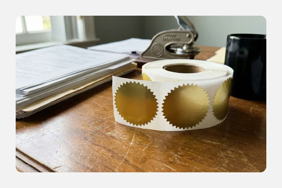 גליל מדבקות נוטריון מוזהבות ומשוננות על שולחן עץ לצד ציוד משרדי משפטי.