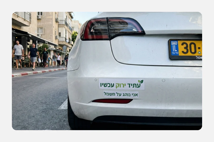 מדבקת פגוש לבנה מויניל PVC על רכב טסלה לבן עם הכיתוב "עתיד ירוק עכשיו" ו"אני נוהג על חשמל" בעברית.