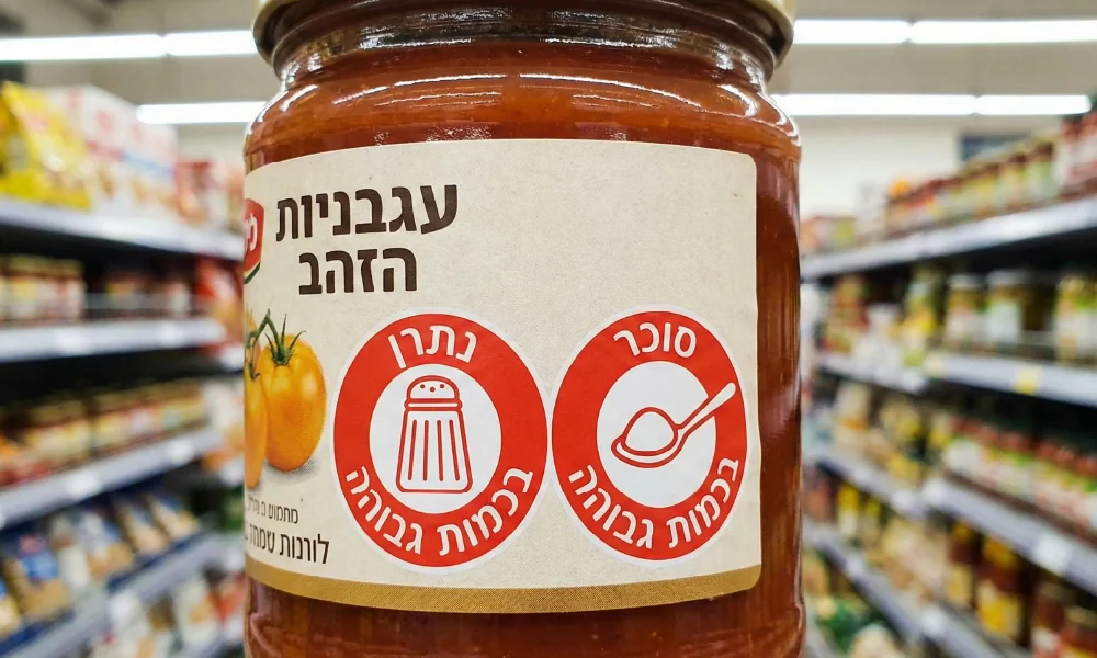צנצנת רוטב עגבניות עם מדבקות אזהרה אדומות של משרד הבריאות המציינות נתרן וסוכר בכמות גבוהה.